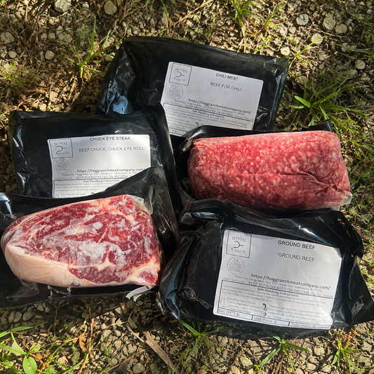 Five vacuum-sealed packages of beef products arranged on gravel outdoors: 2 chuck eye steaks, 2 ground meat packs, and a black packages labeled chili beef