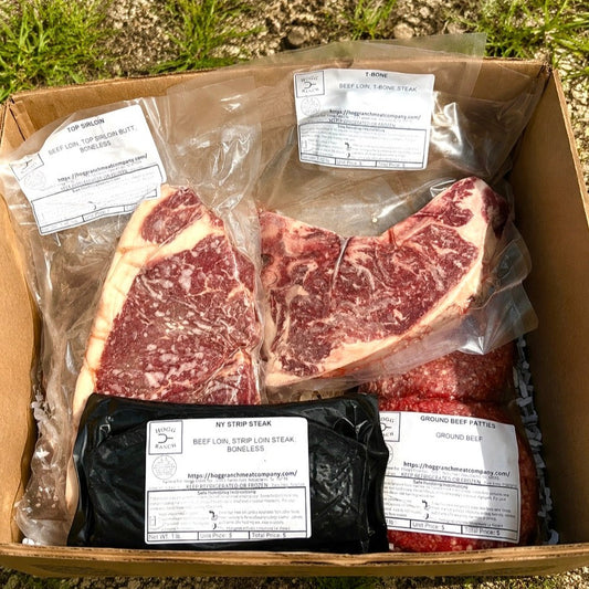 A cardboard shipping box containing packaged beef sirloin & NY strip steaks and beef patties, photographed outdoors on gravel and grass.
